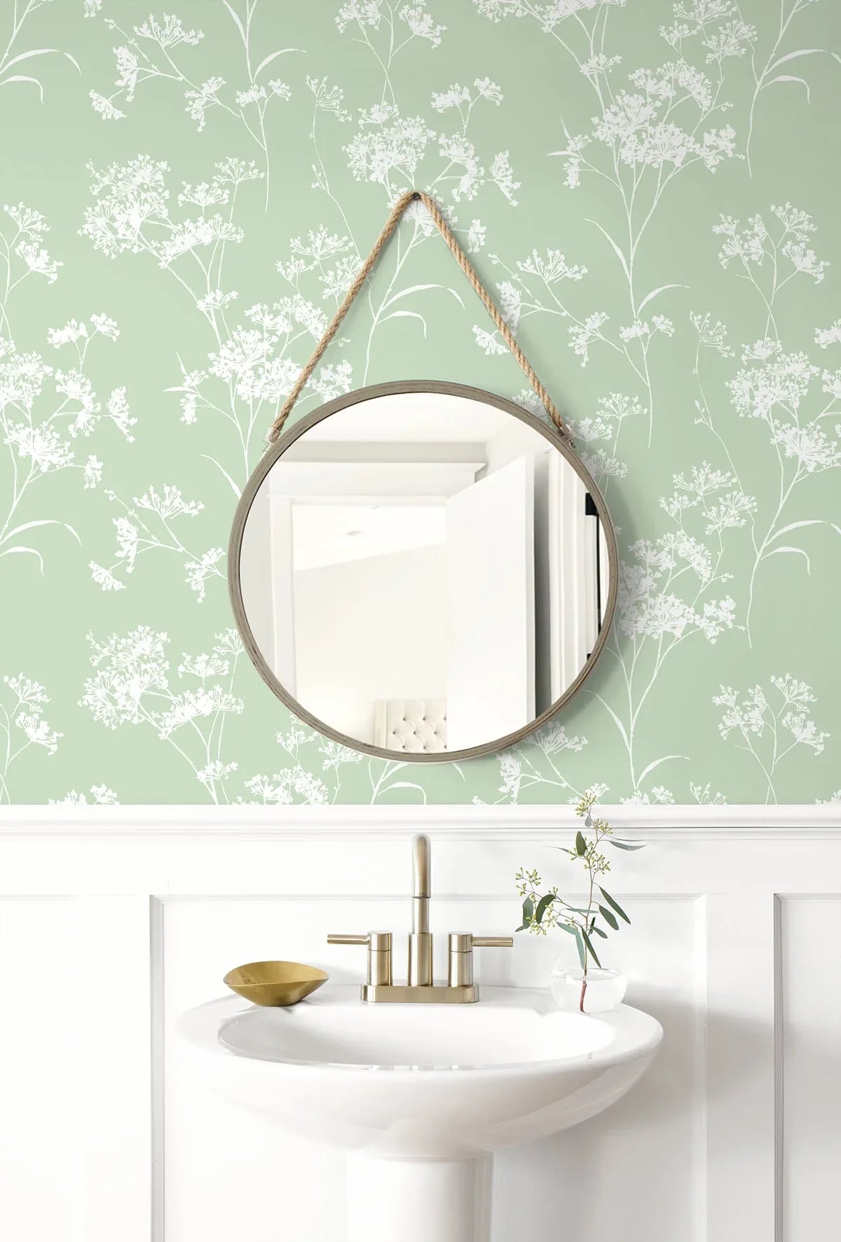 Round mirror with rope hangs above sink, set against Soft Scent Floral Peel Stick wallpaper and white wainscoting.
