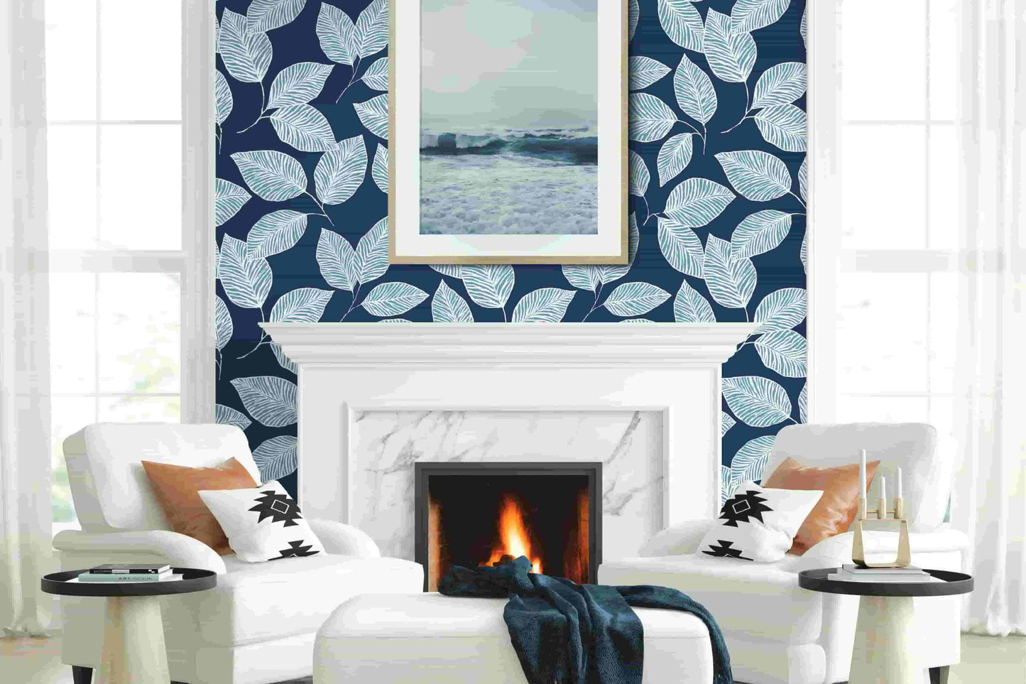 White armchairs by a marble fireplace with fire, Stylized Foliage Wallpaper feature wall, and framed ocean artwork above.