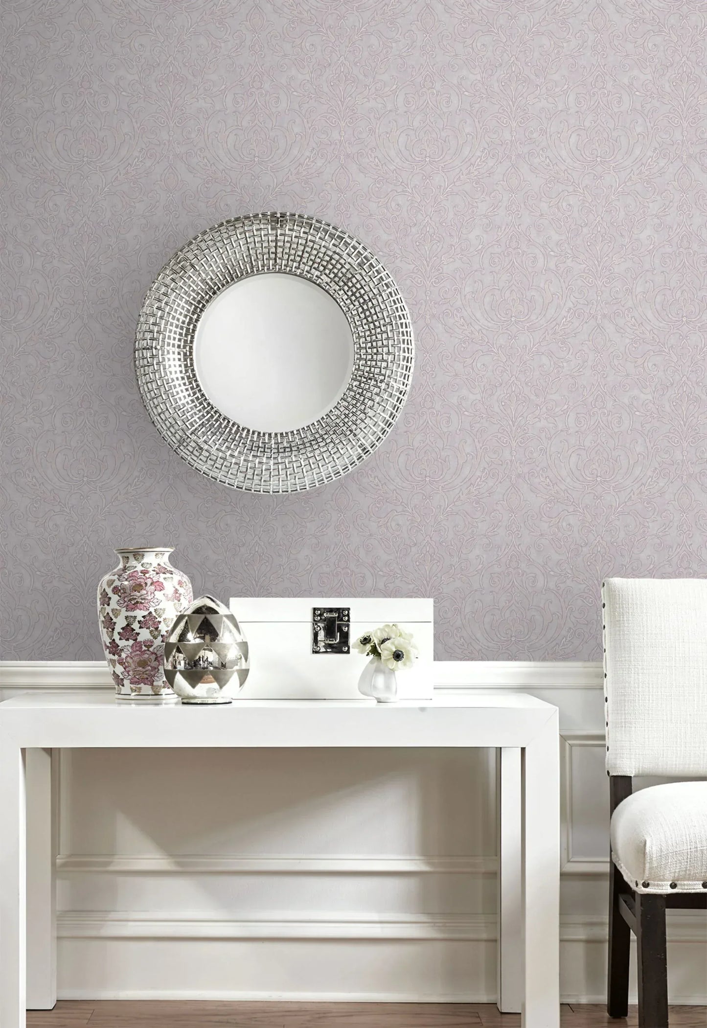 Round silver mirror above a white table with vases, flowers, and chair against Soft Damask Wallpaper.