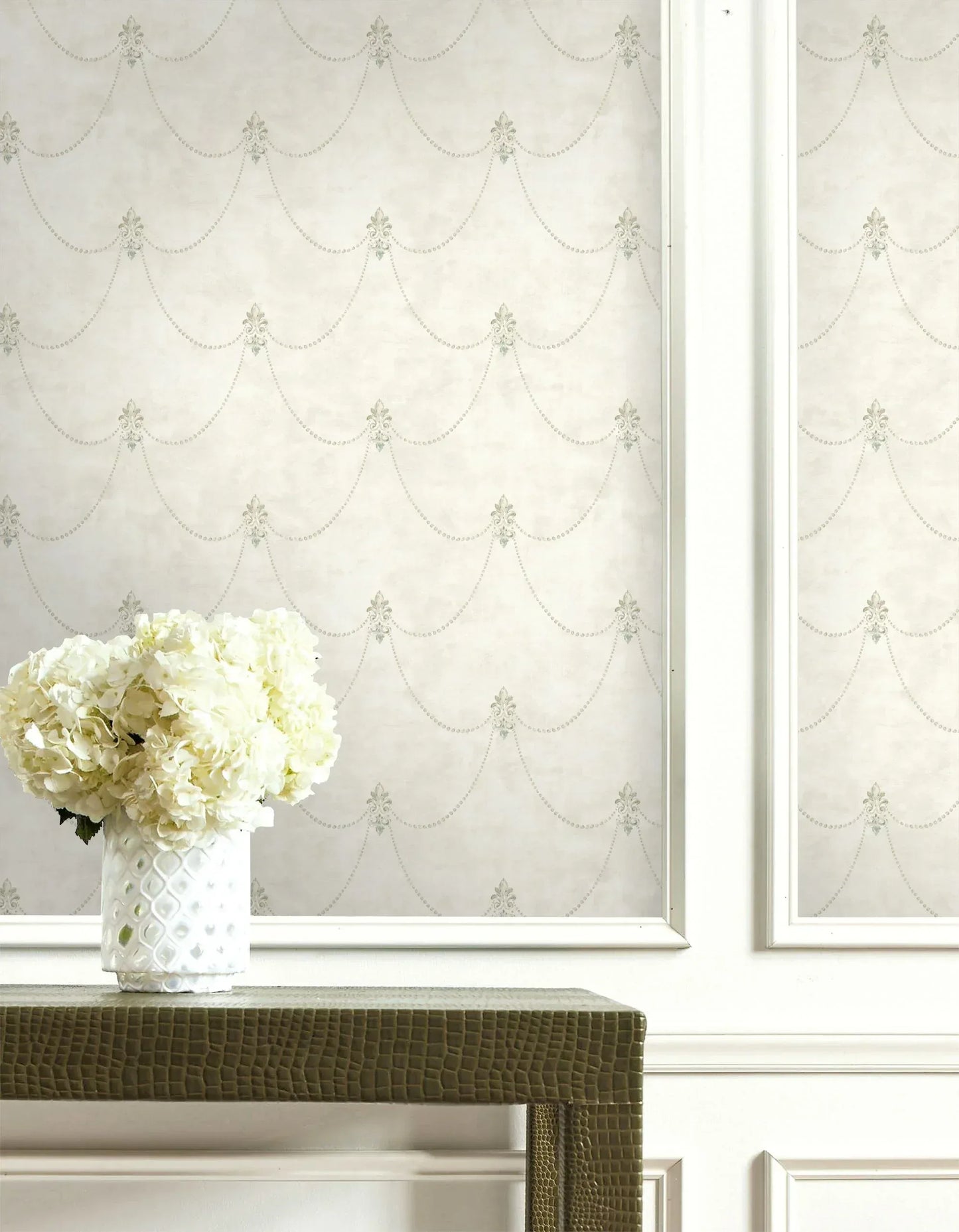 White hydrangeas in a textured vase sit on a green table before cream Fleur de Lys Wallpaper.