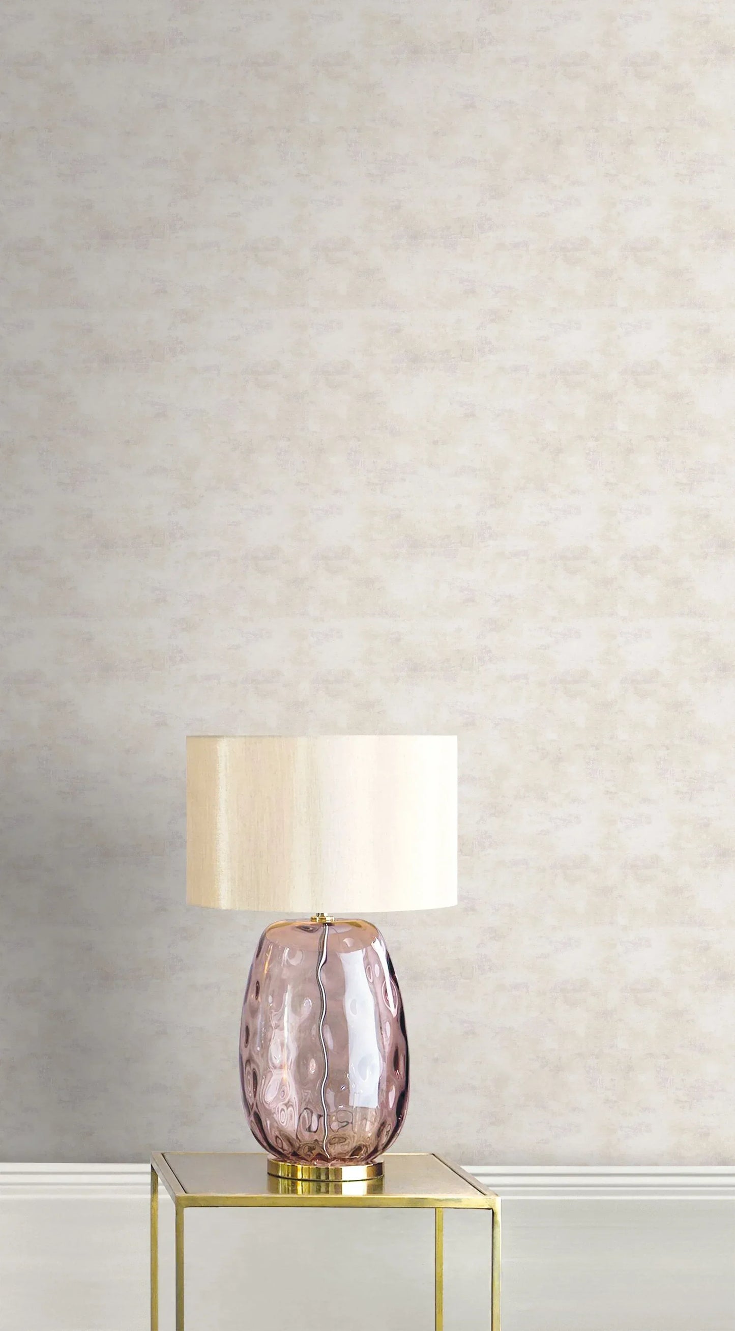 A pink glass table lamp with a beige shade sits on a gold table against Stucco Faux Wallpaper.