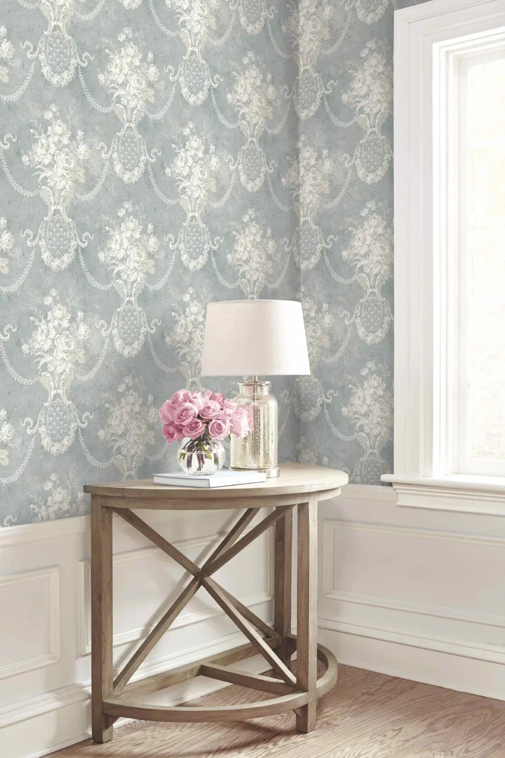 A small wooden table with a lamp and pink flowers beside a window, against Cameo Spatula Wallpaper.