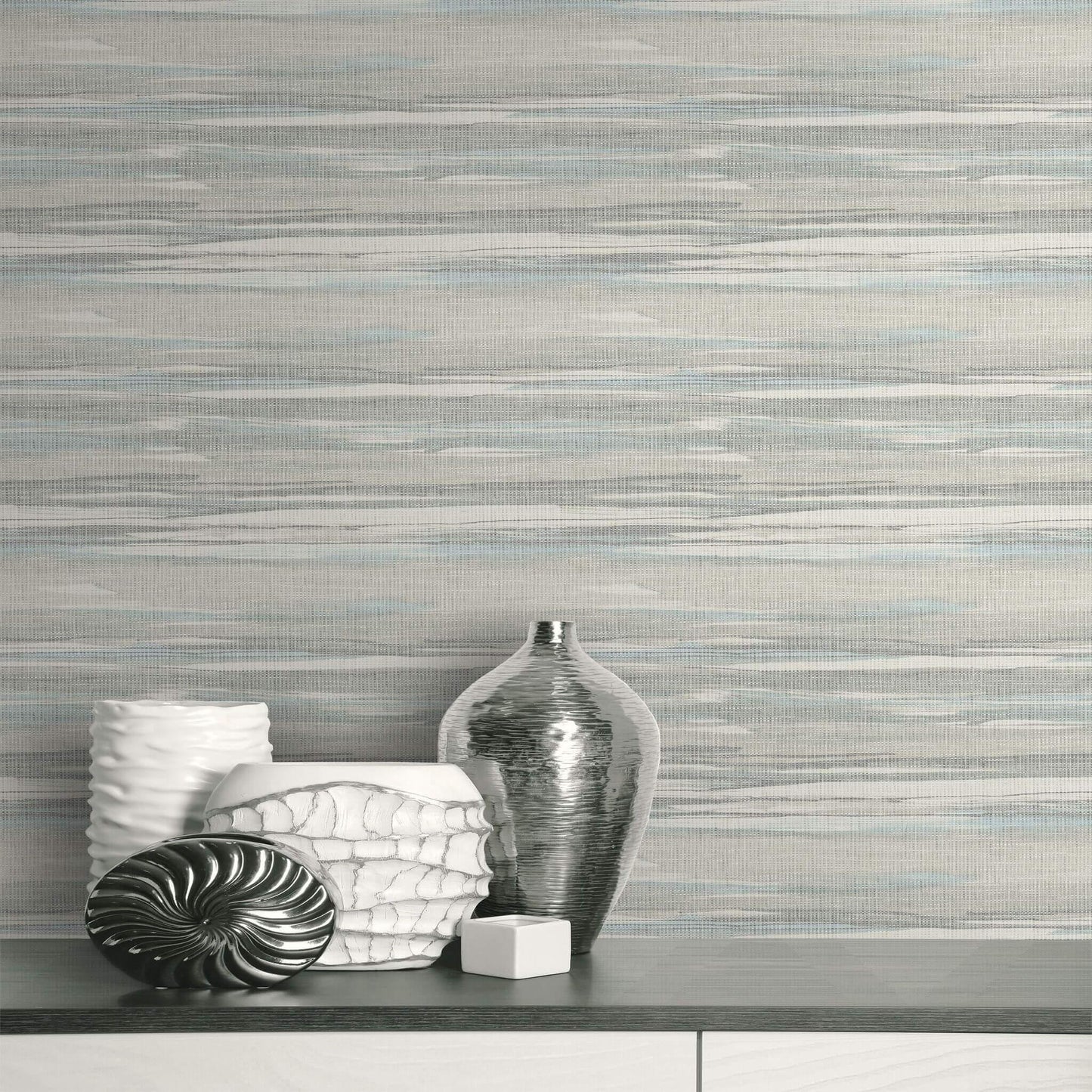 Silver and white textured vases on a gray surface with Grasscloth Waves Wallpaper in soft gray tones behind.