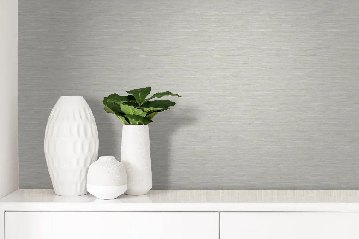 Three white vases, one holding a green plant, sit on a white cabinet against metallic texture wallpaper in light gray.