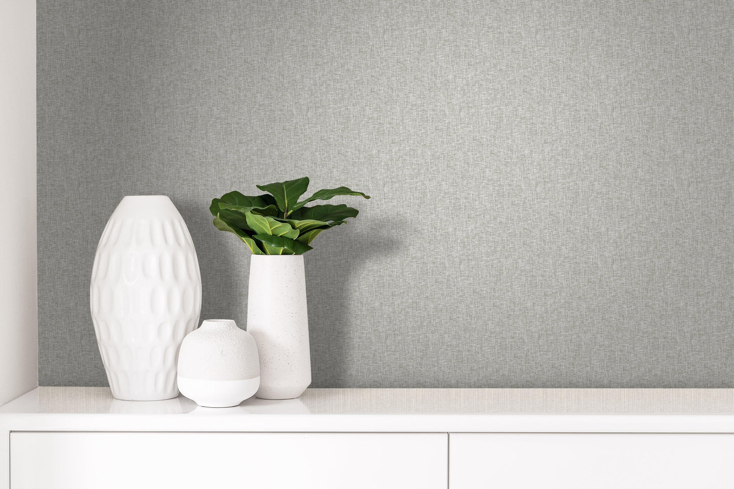 Three white vases, one with green leaves, on a white cabinet against Textile Plain Linen Wallpaper.