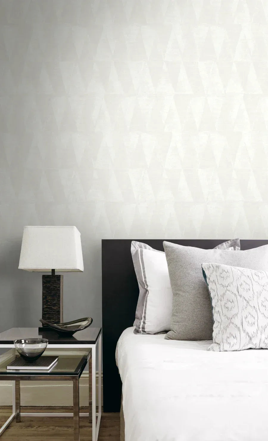 Minimalist bedroom with Small Triangles Wallpaper, a modern lamp, glass side table, and neatly arranged gray and white pillows.