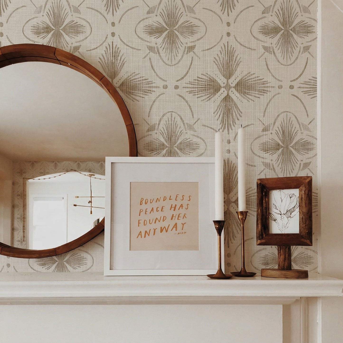 A round mirror, framed art, and candlesticks sit on a mantel against cream Tropical Bali Essence grasscloth wallpaper.
