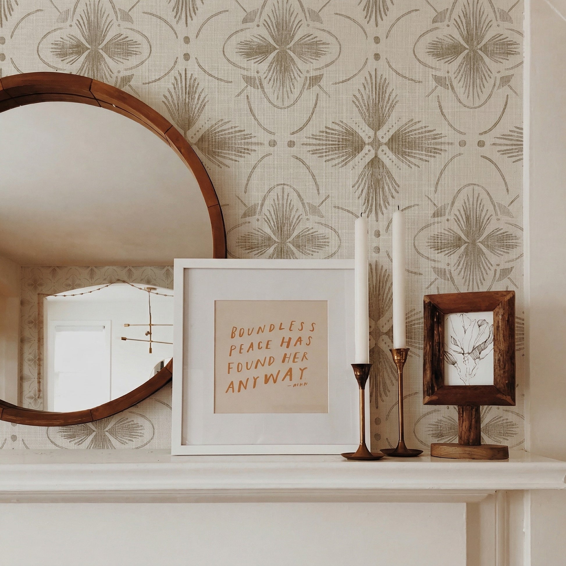 A round mirror, framed art, and candlesticks sit on a mantel against cream Tropical Bali Essence grasscloth wallpaper.
