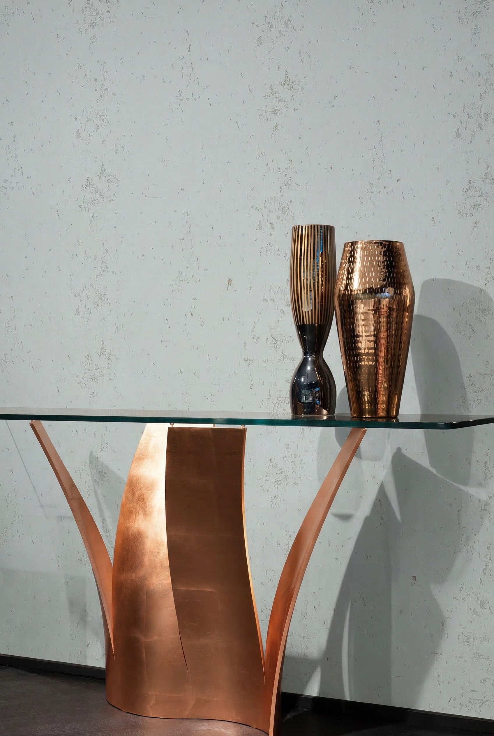 A glass table with a sculptural copper base holds two metallic vases before Algarve Cork Grasscloth Naturals.