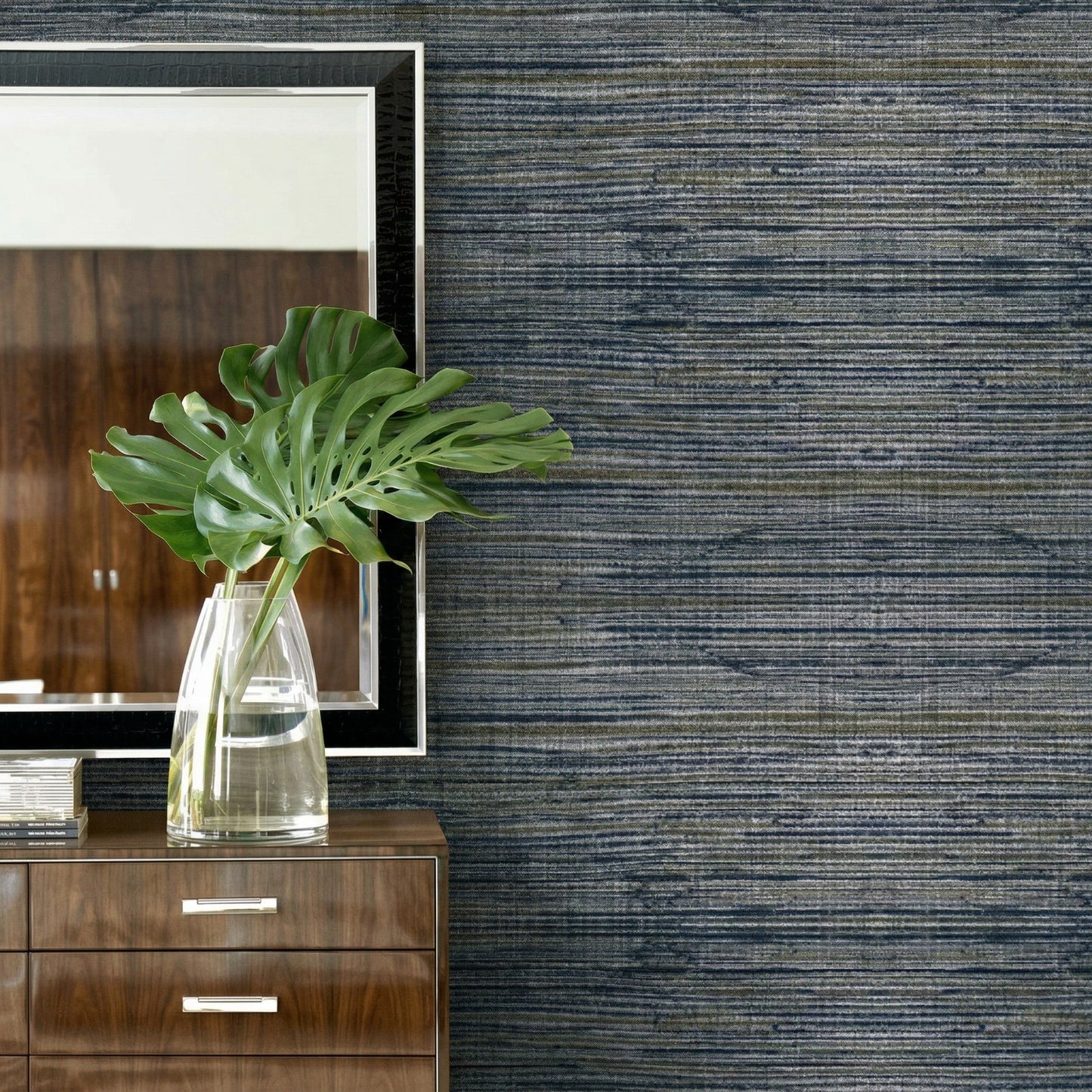 A glass vase with green leaves sits on a dresser beside a mirror, set against Straw Texture Grasscloth Naturals wallpaper.