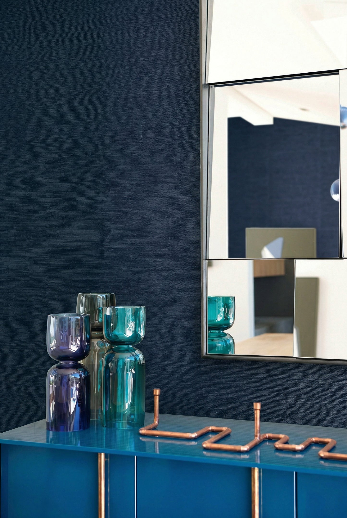 A blue sideboard with glass vases stands by a wall mirror against Sisal Texture Grasscloth Naturals wallpaper.