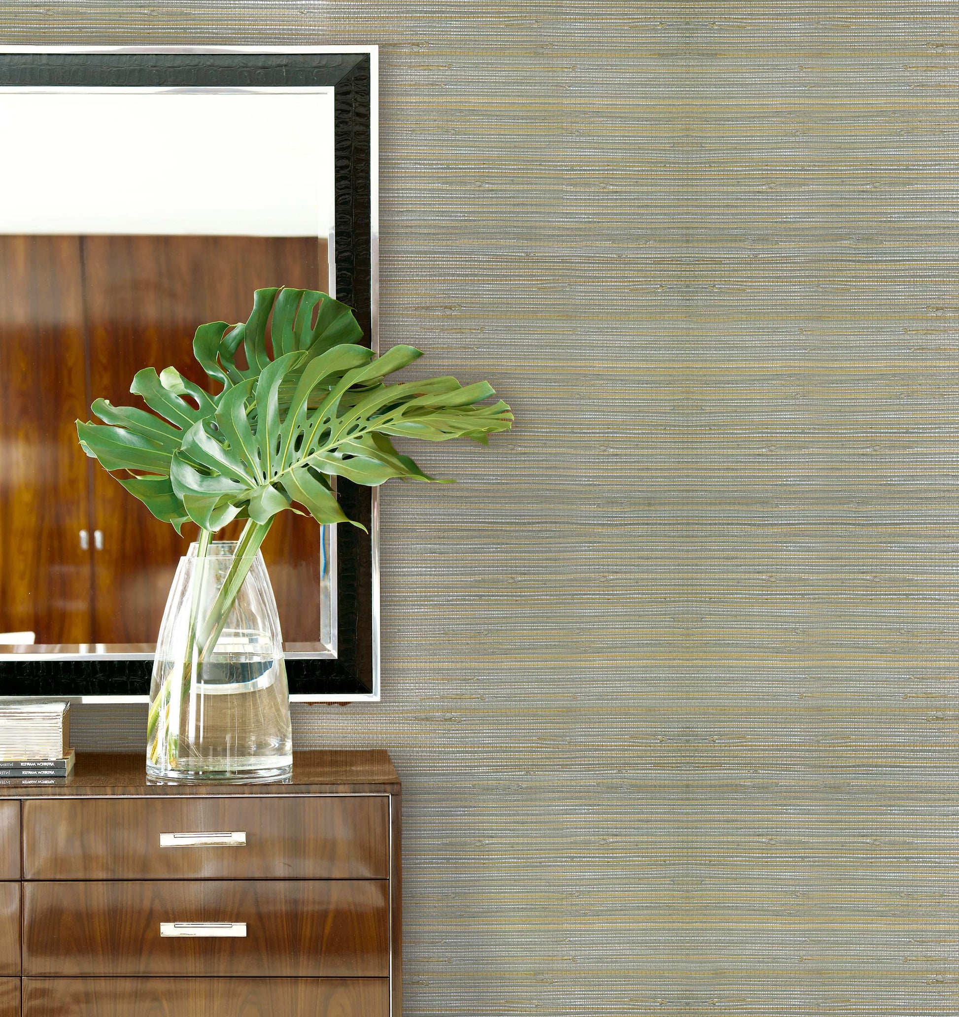 A glass vase with large green leaves sits on a wooden cabinet beside a mirror on Straw Texture Grasscloth.