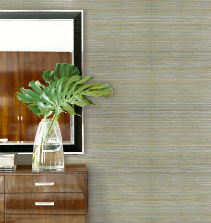 A glass vase with large green leaves sits on a wooden cabinet beside a mirror on Straw Texture Grasscloth.