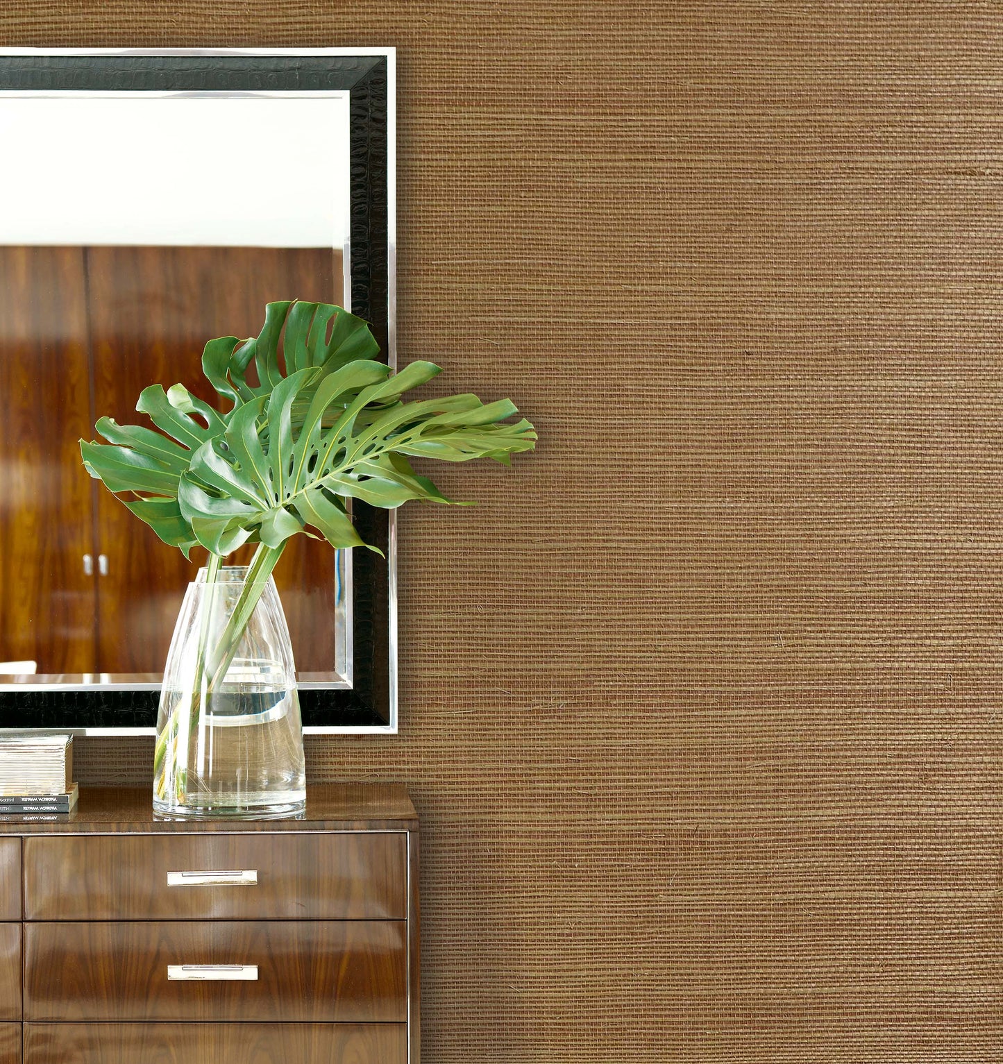 A glass vase with green monstera leaves sits on a wooden dresser against Sisal Texture Grasscloth Naturals wallpaper.