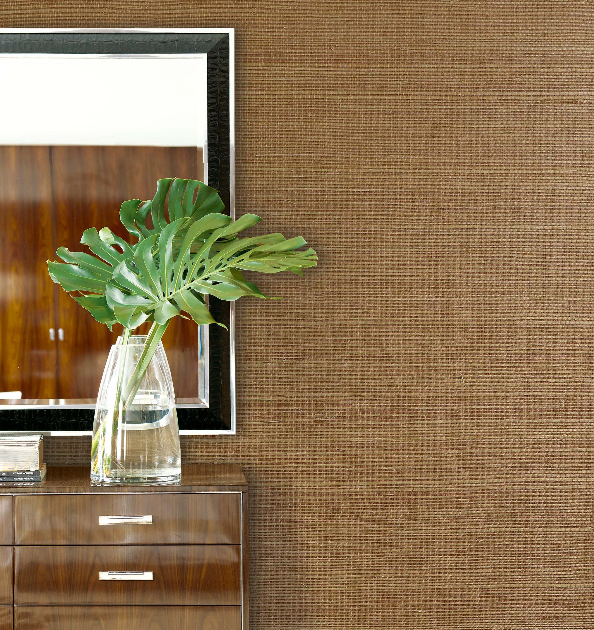 A glass vase with green monstera leaves sits on a wooden dresser against Sisal Texture Grasscloth Naturals wallpaper.