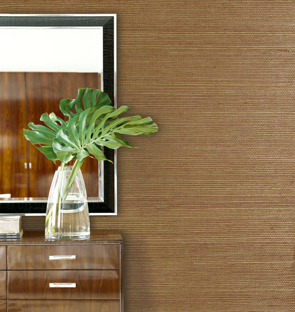 A glass vase with green monstera leaves sits on a wooden dresser against Sisal Texture Grasscloth Naturals wallpaper.
