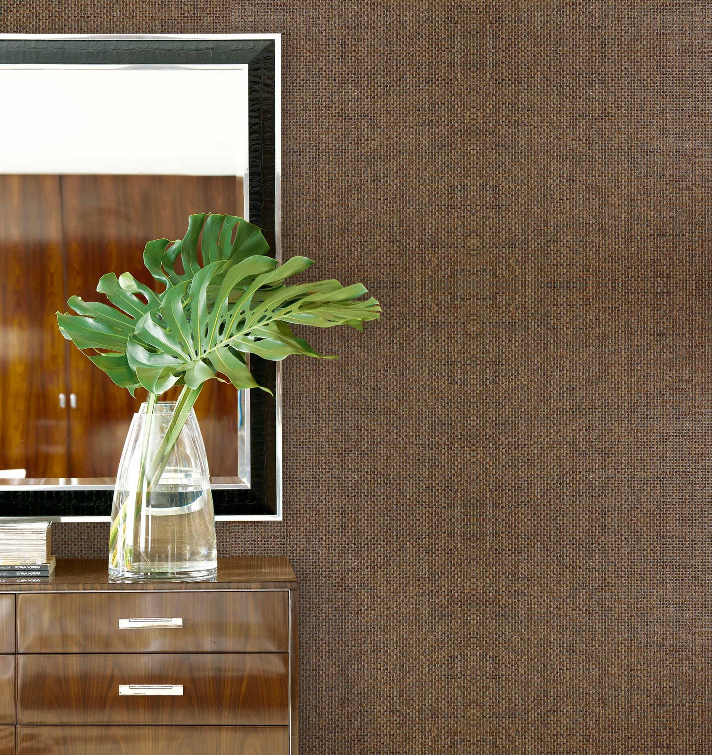 A glass vase with green leaves sits on a dresser beside a mirror, against Paperbraid Texture Grasscloth Naturals wallpaper.