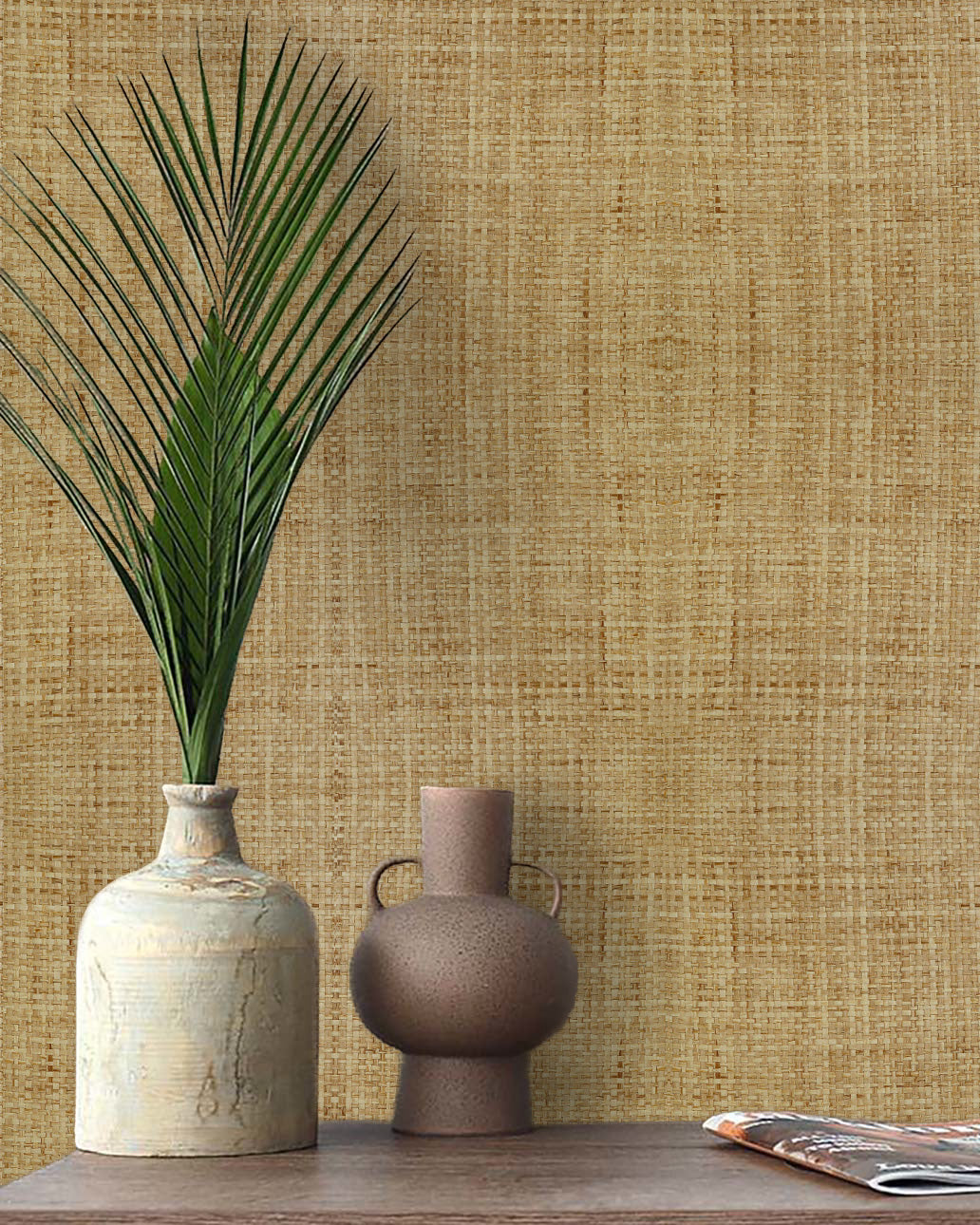 A palm leaf in a ceramic vase sits on wood with Paper Interlace Texture Grasscloth Naturals wallpaper behind.