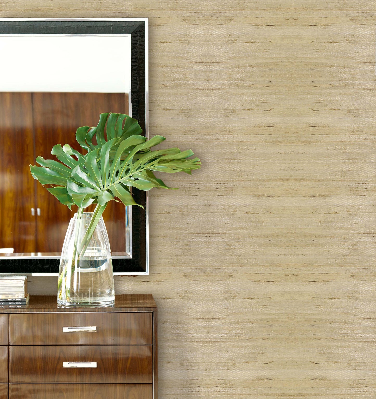 A vase with monstera leaves sits on a wooden dresser beside a mirror and Straw Texture Grasscloth Naturals wallcovering.
