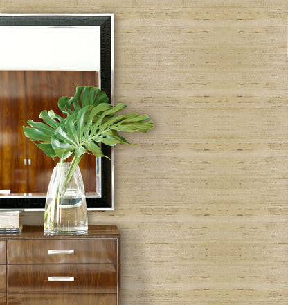 A vase with monstera leaves sits on a wooden dresser beside a mirror and Straw Texture Grasscloth Naturals wallcovering.