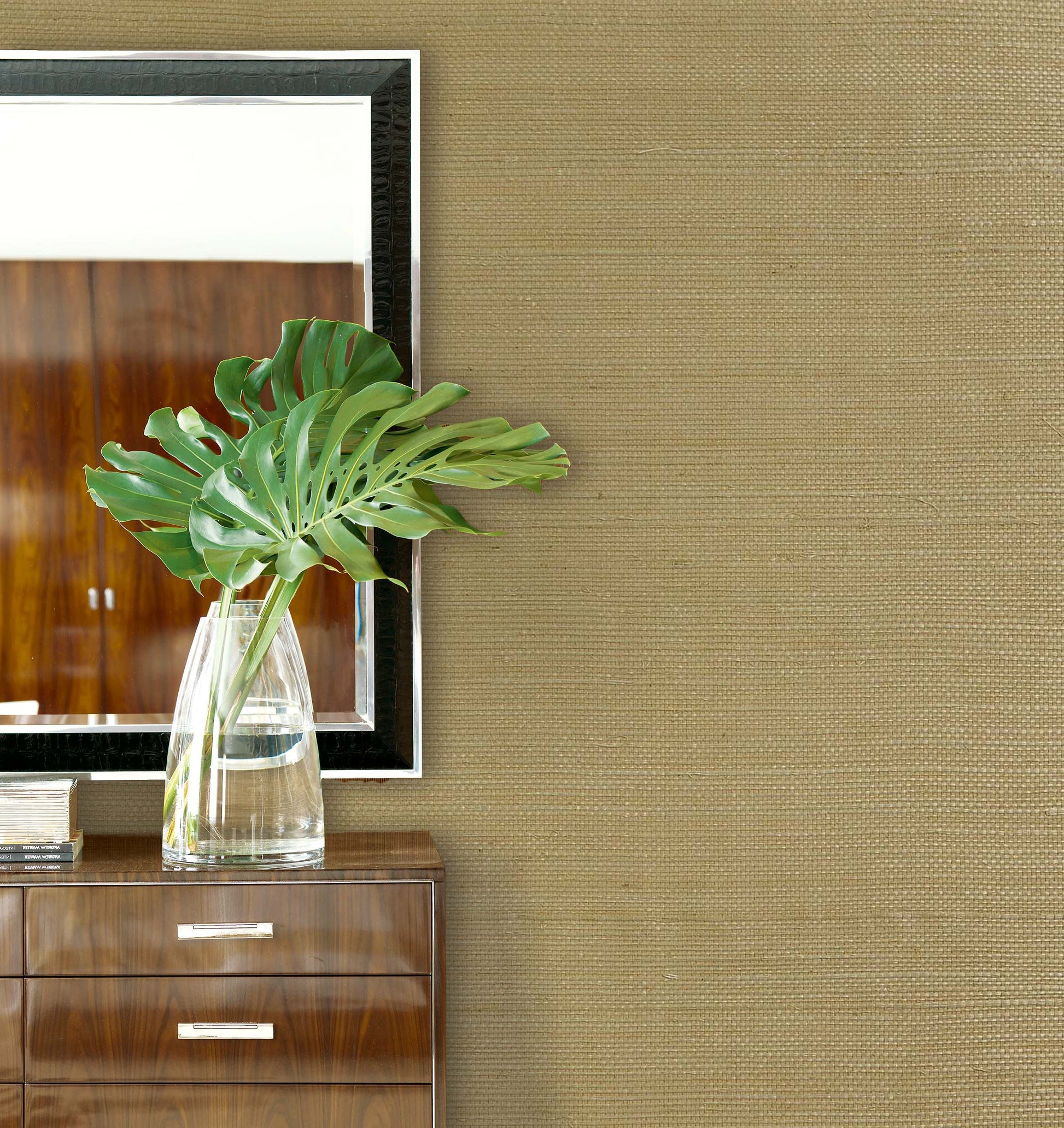 Large green leaves in a clear vase sit on a dresser below a mirror with Sisal Texture Grasscloth Naturals wallpaper.