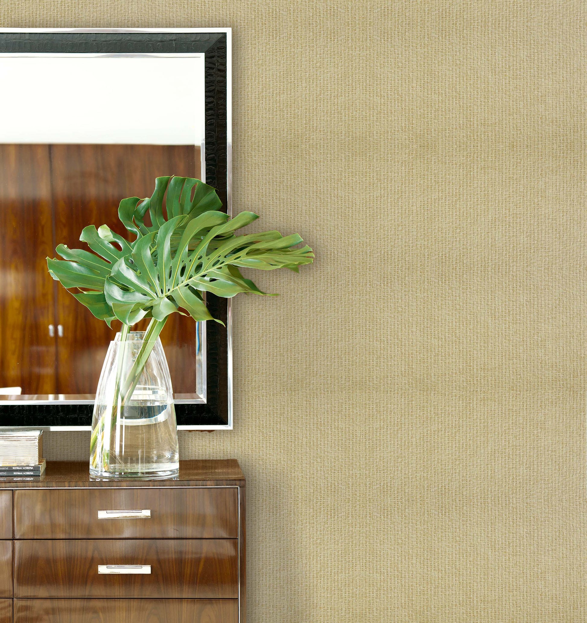 A glass vase with green leaves on a wooden dresser, beside a mirror and Paperbraid Texture Grasscloth Naturals wallpaper.