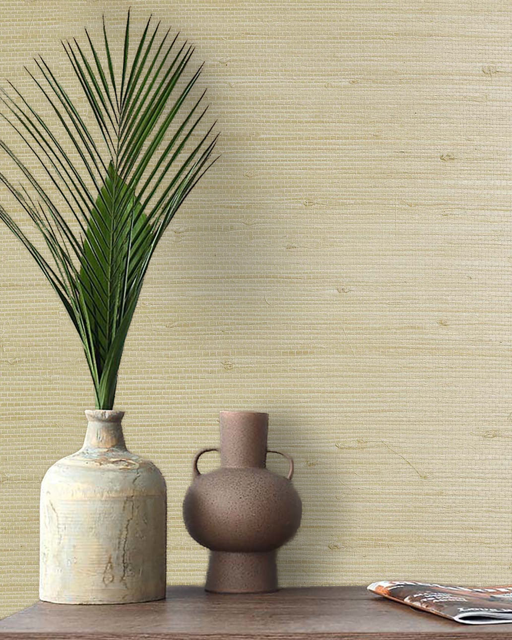 A palm leaf in a ceramic vase and a brown jug on a wooden table with Straw Texture Grasscloth Naturals wall decor.