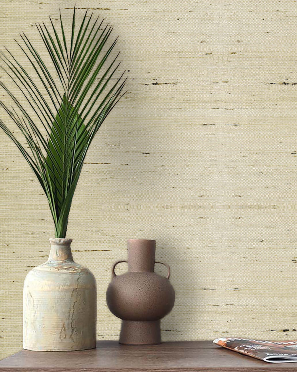 A vase with a palm leaf and another vase sit by a magazine against Straw Texture Grasscloth Naturals wallpaper.