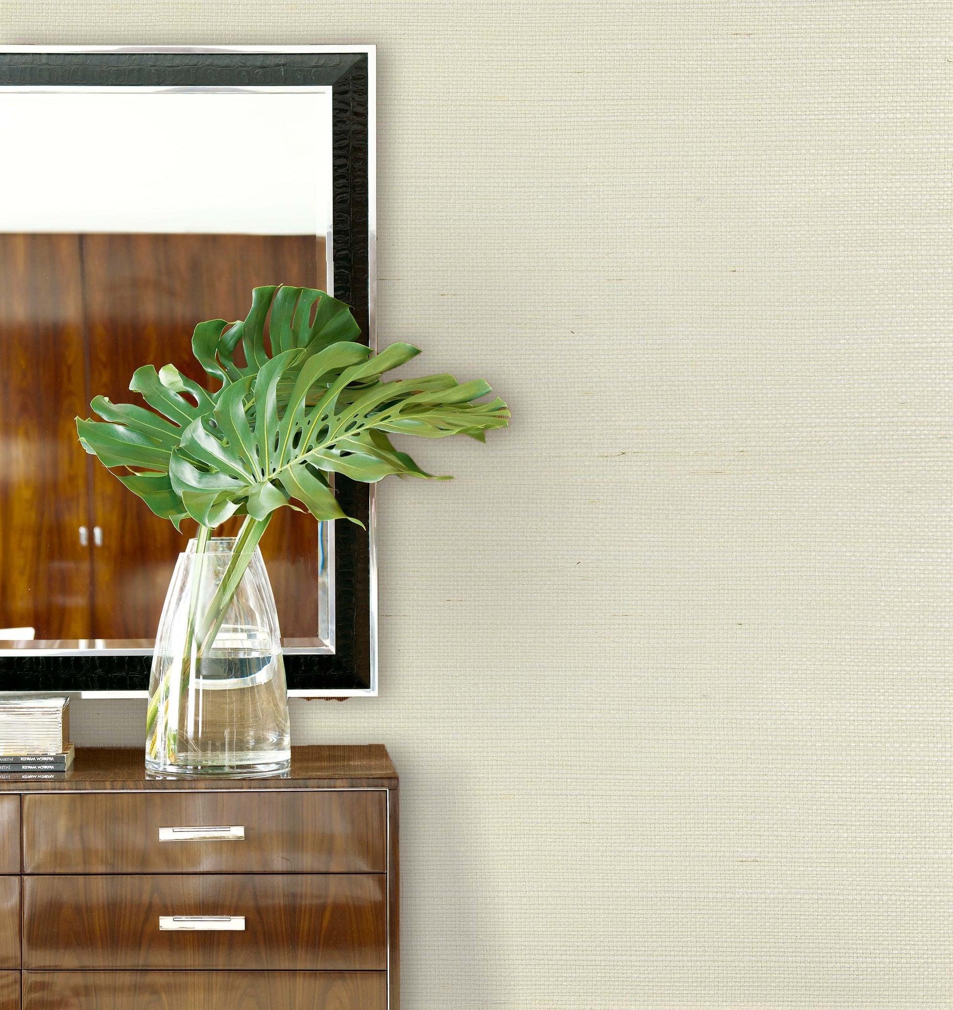 A glass vase with large green leaves sits on a dresser against Sisal Texture Grasscloth Naturals wallpaper and a mirror.