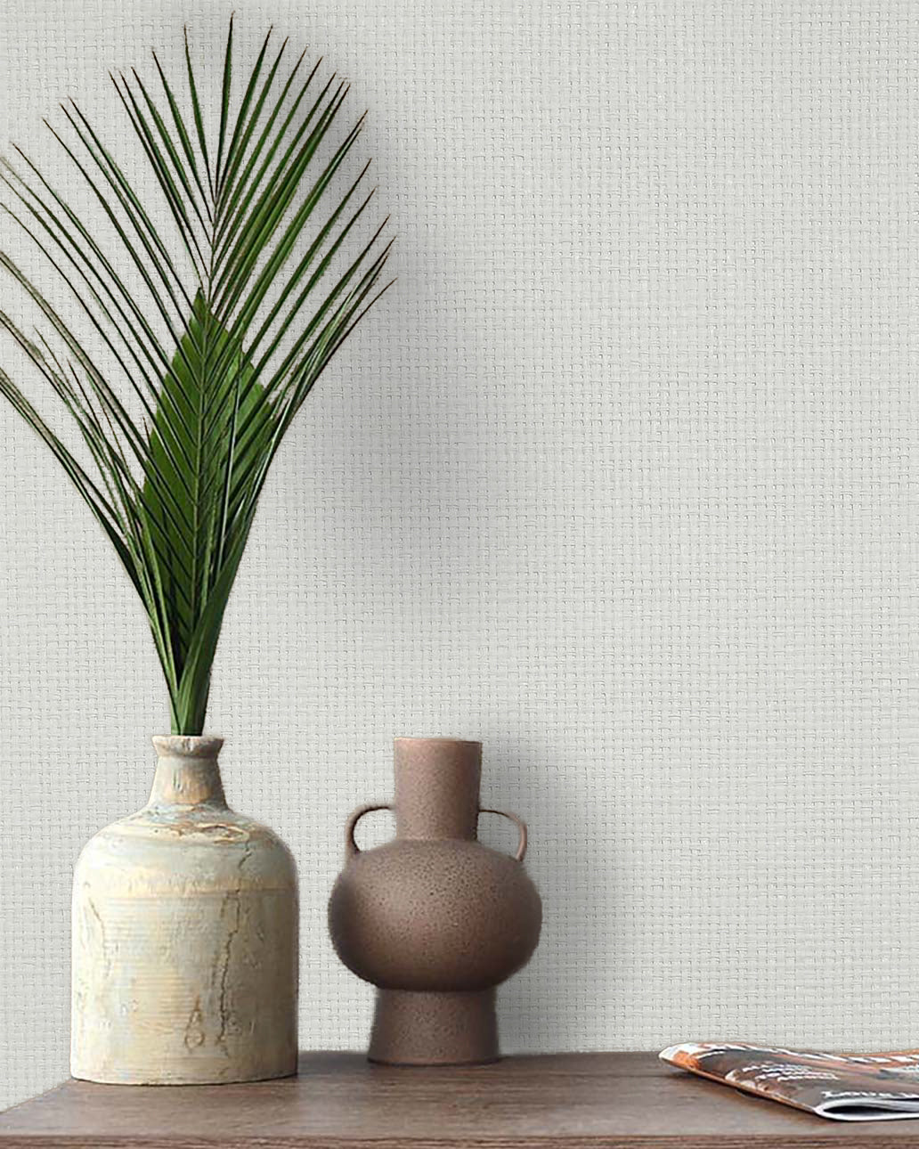 Two ceramic vases, one with a palm frond, sit by a magazine and Paperbraid Texture Grasscloth Naturals wallpaper.