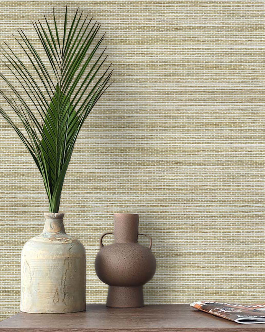 A ceramic vase with palm fronds and a small jug sit on a wooden table against Straw Texture Grasscloth Naturals.