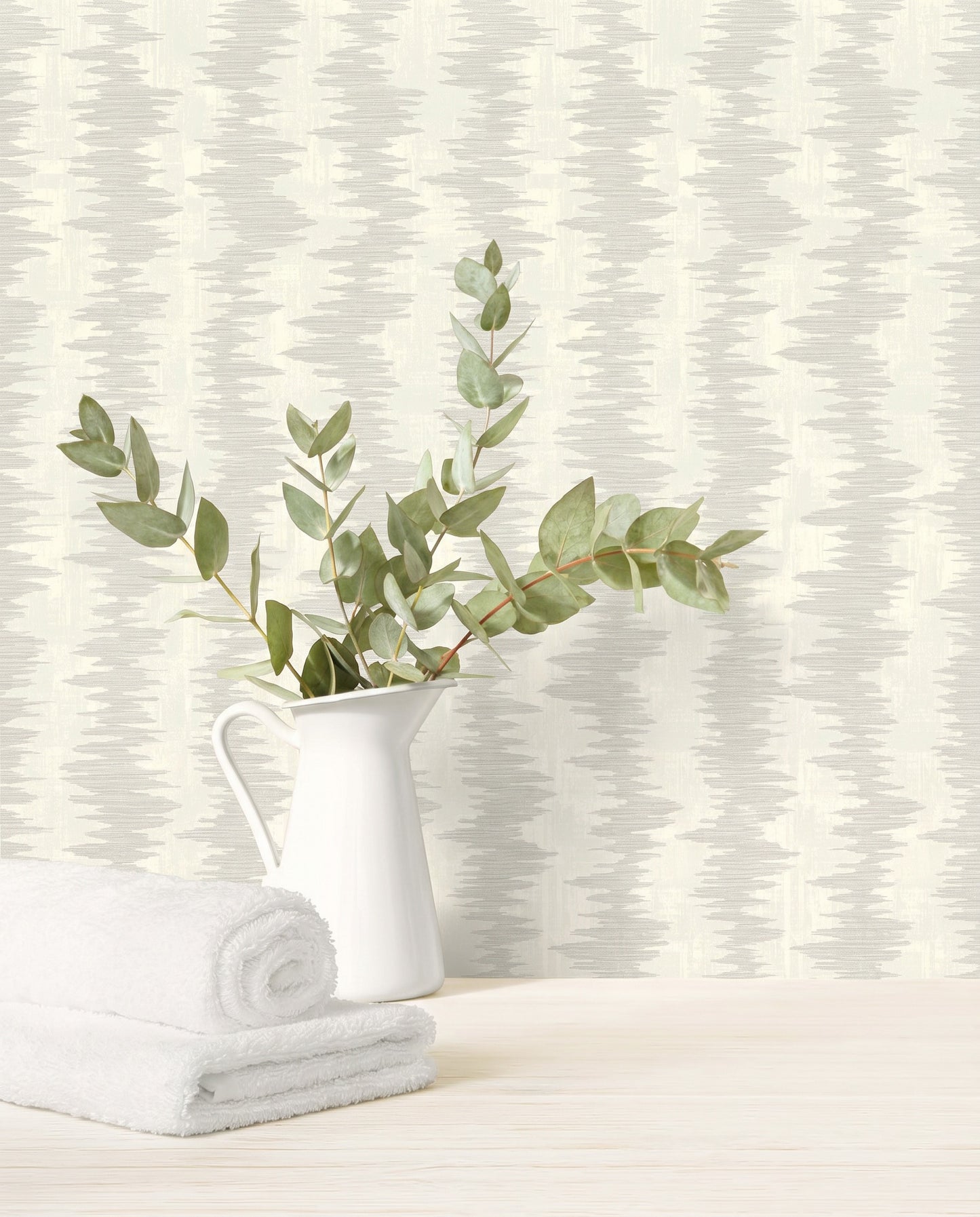 White pitcher with leafy branches and towels on wood, set against Ikat Stripe Wallpaper in the background.