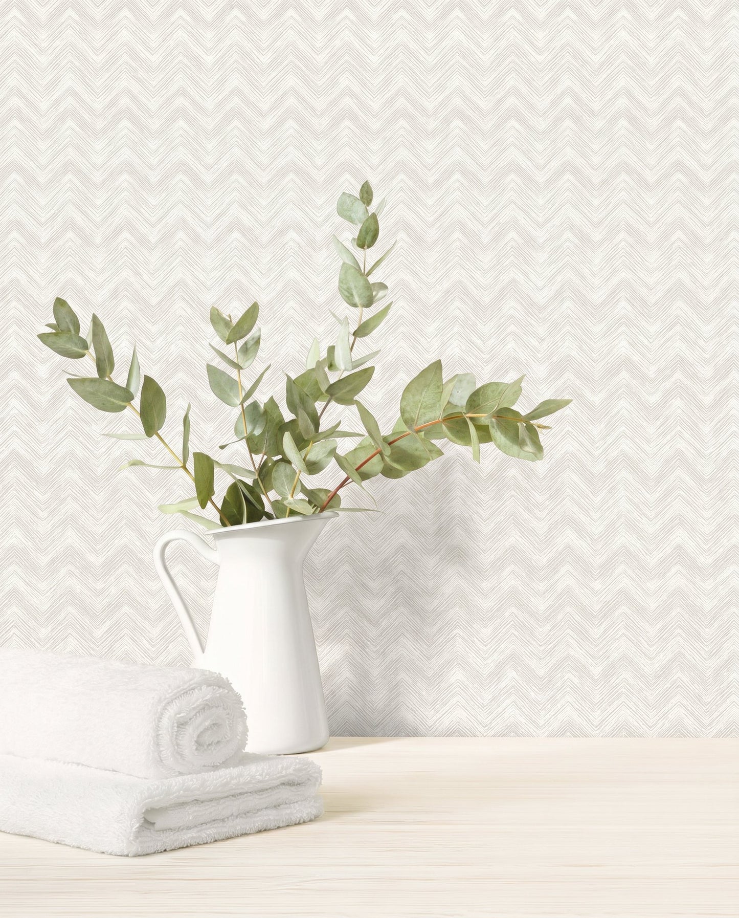 White pitcher with eucalyptus branches and towels on wood surface, set against Chevron Wallpaper in white/off white.