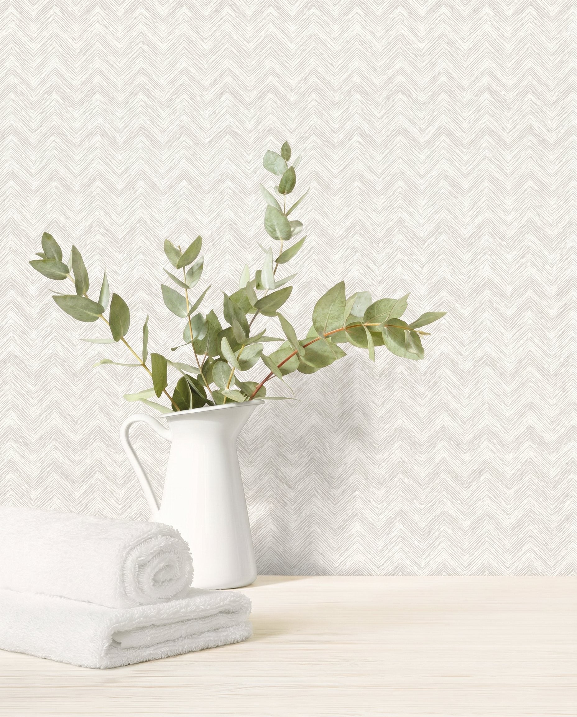 White pitcher with eucalyptus branches and towels on wood surface, set against Chevron Wallpaper in white/off white.