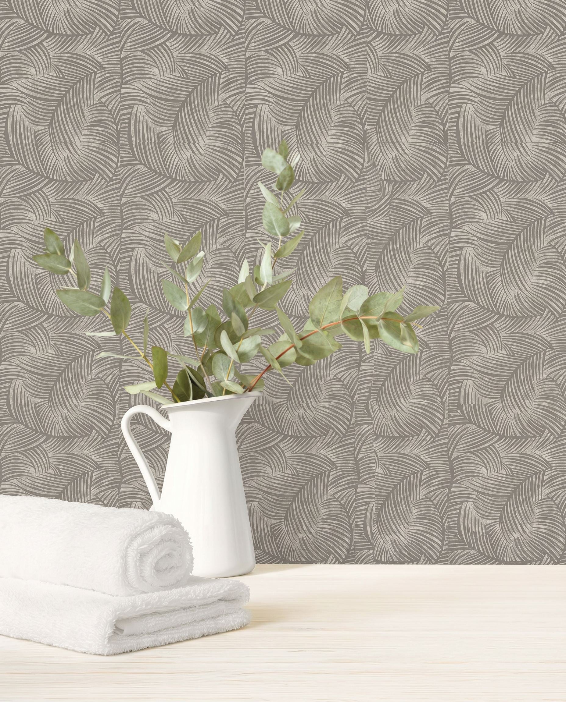 A white pitcher with leafy branches and towels rests on a light surface, set against Palm Fronds Wallpaper.