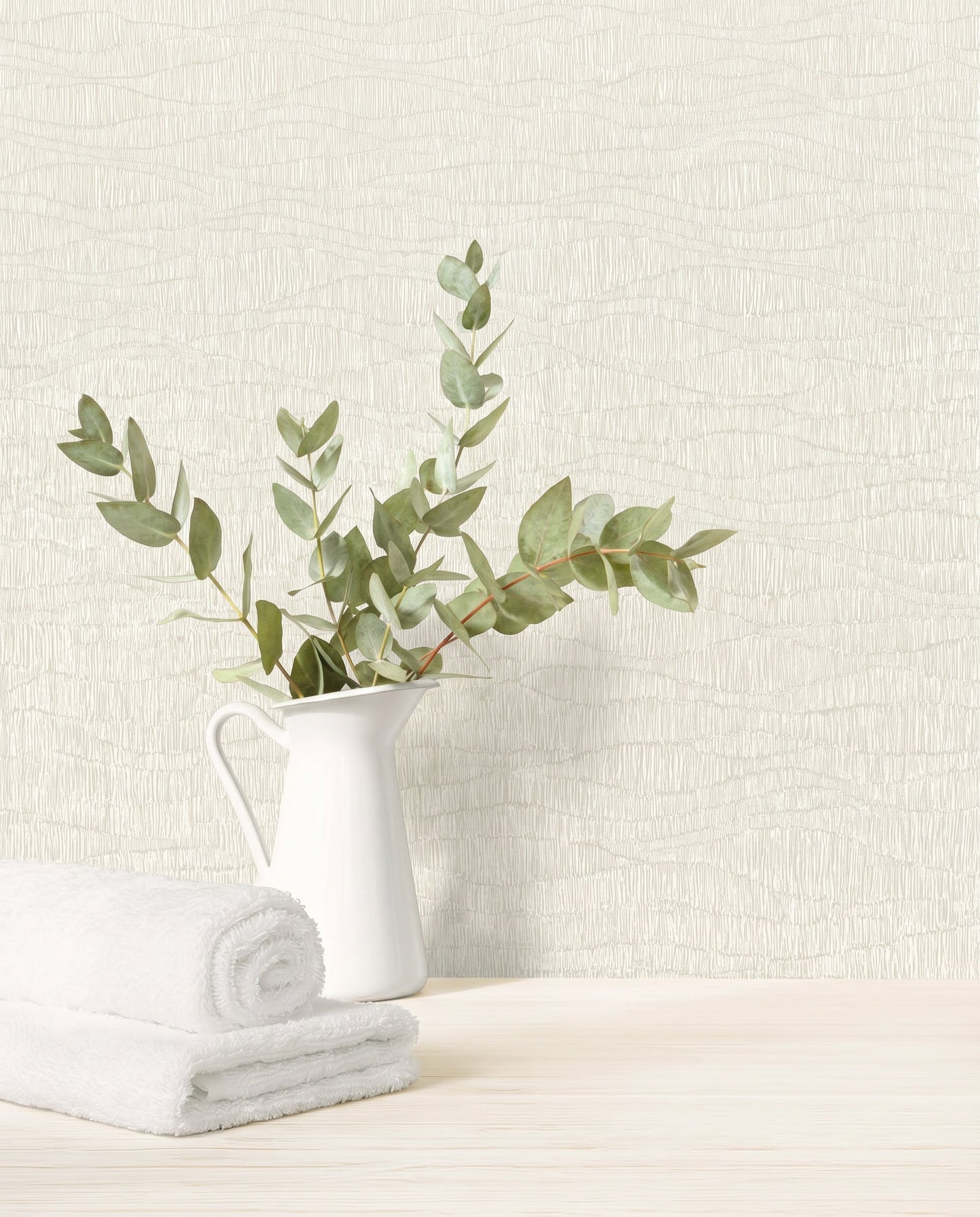 A white pitcher with green eucalyptus beside rolled towels on a light wood surface with Wavy Wallpaper.