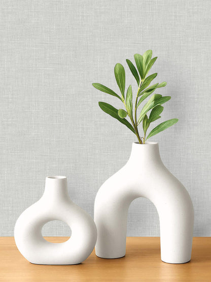 Two modern white ceramic vases, one with leafy stems, on wood against washable Faux Linen Wallpaper in light gray.