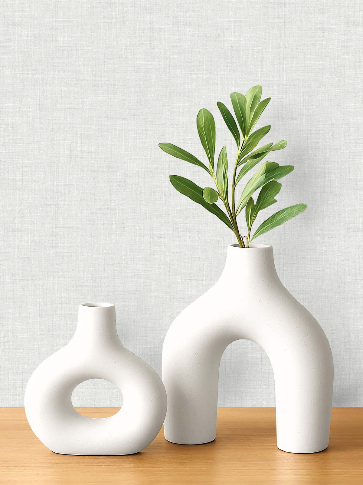 Two modern white ceramic vases with green leaves sit on a wooden surface against faux linen wallpaper.