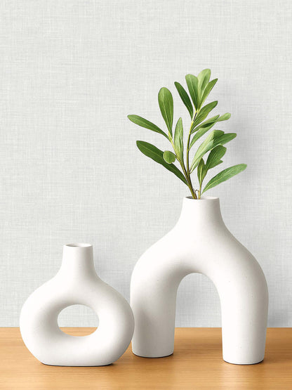 Two modern white ceramic vases with green leaves sit on a wooden surface against faux linen wallpaper.