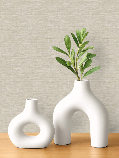 Two modern white ceramic vases sit on wood, set against Pearly Linen Wallpaper with a textured beige finish.