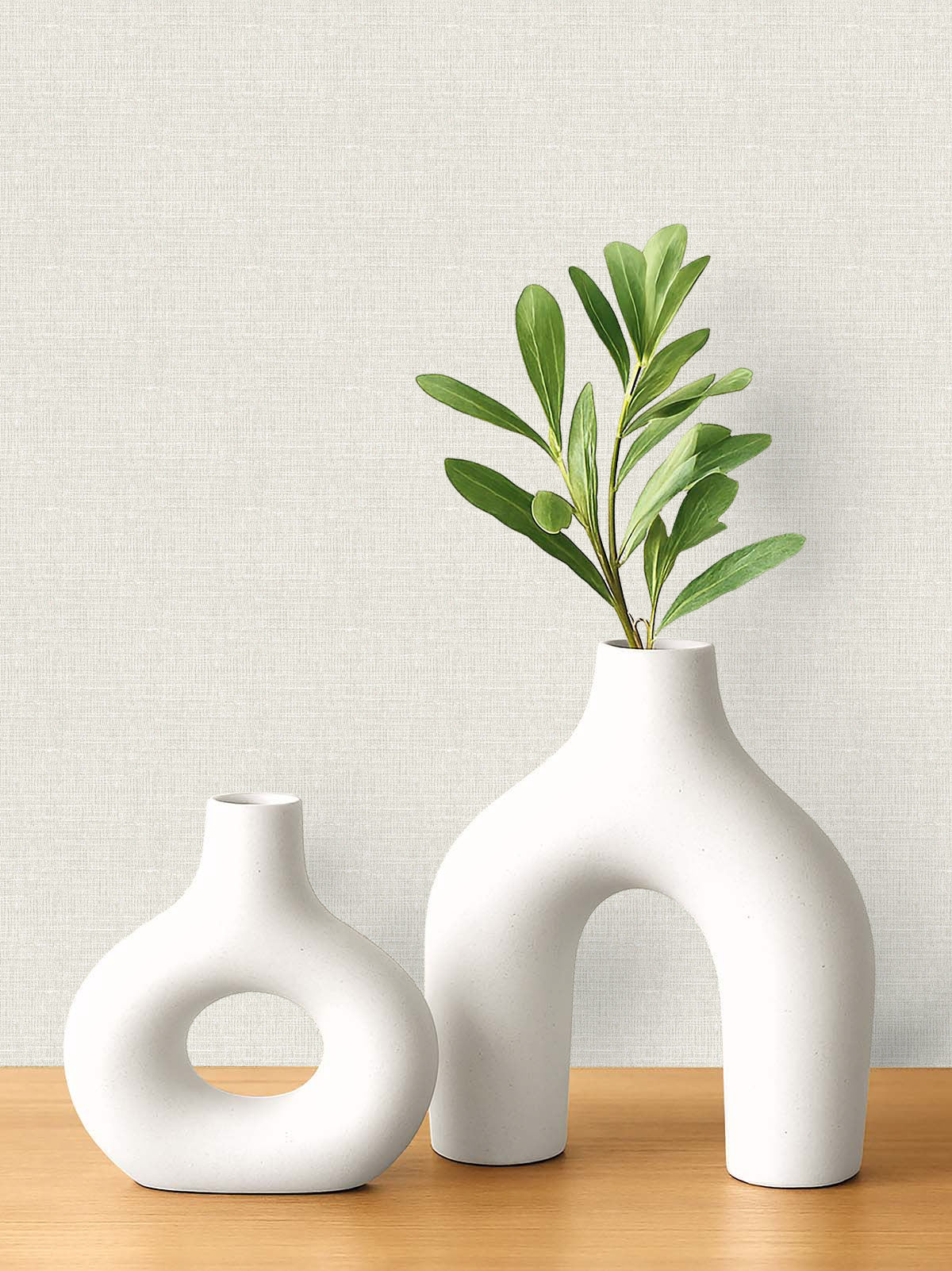 Two white ceramic vases with leafy branches on wood, set against Woven Linen Effect Wallpaper in light tones.