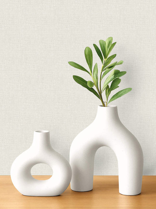 Two white ceramic vases with leafy branches on wood, set against Woven Linen Effect Wallpaper in light tones.