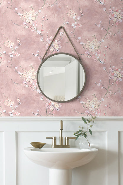 Round mirror on a wall decorated with Pink Blossom Branch Peel & Stick Casa Mia Wallcovering. above a white sink with gold fixtures.