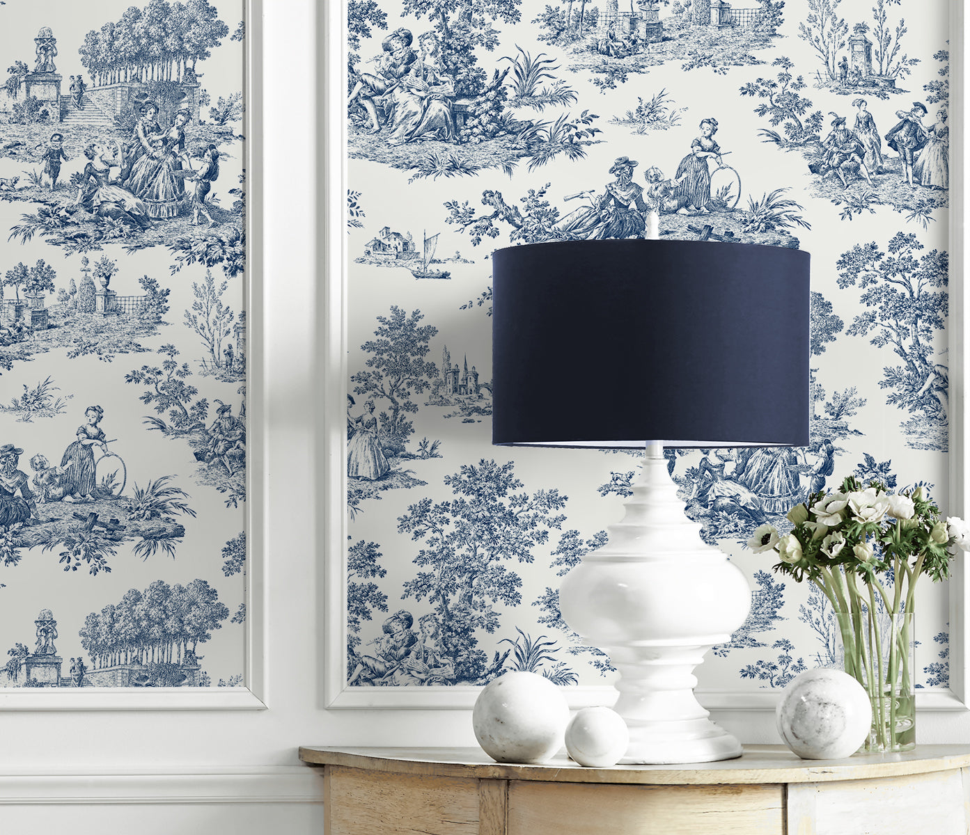 White table with a white lamp, decorative balls, and a vase of flowers against a wall decorated with Blue Toile de Jouy Peel & Stick Casa Mia Wallcovering.