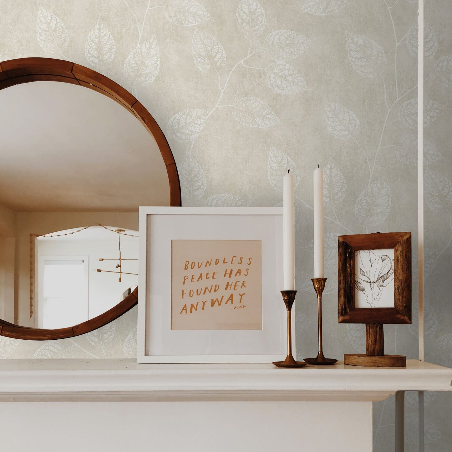Decorative setup with a mirror, framed text, and candles on a shelf against a wall decorated with Beige Branch Of Leaves Wallpaper.
