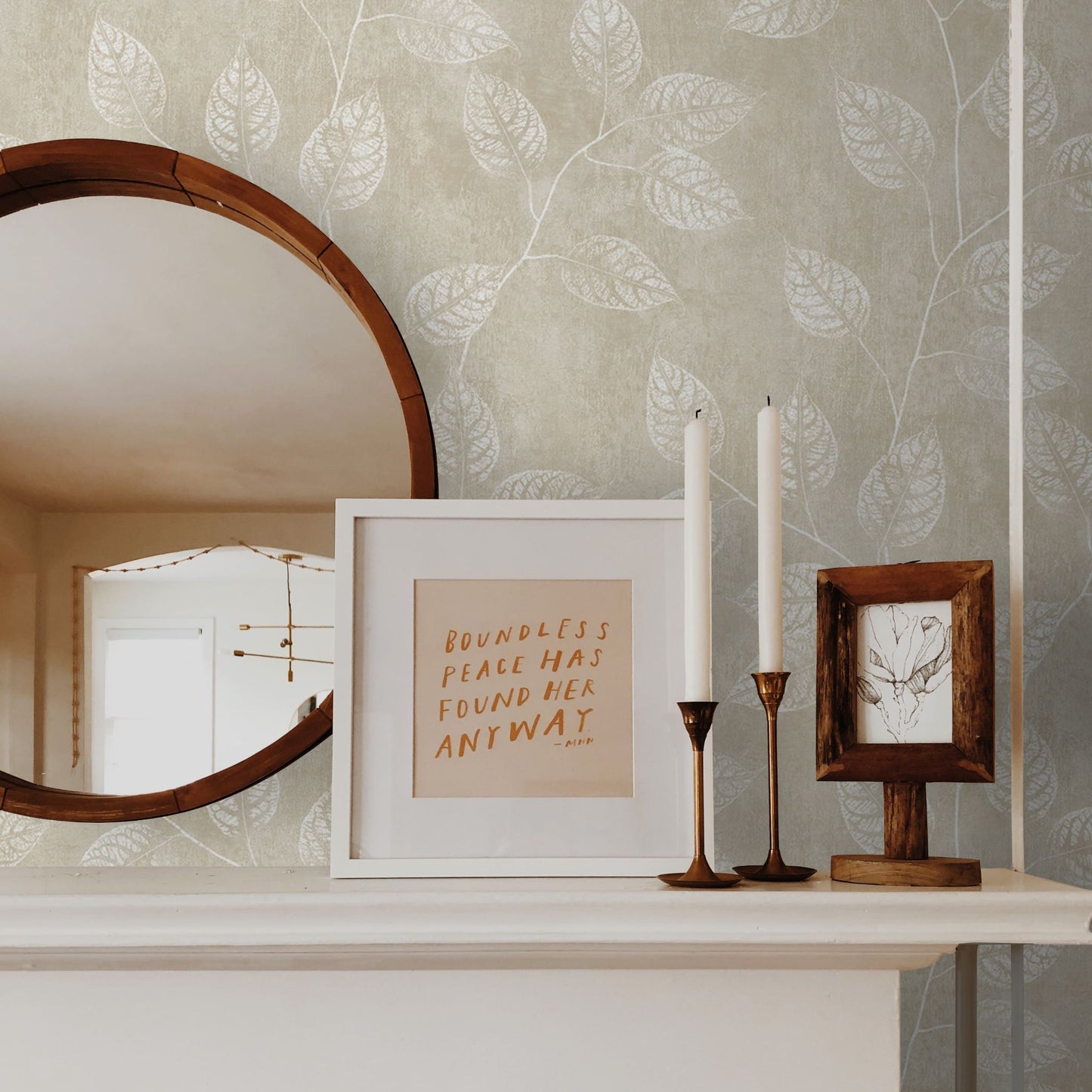Decorative shelf with a mirror, framed quote, candles, and picture frame against a wall decorated with Sand Yellow & Cream Branch Of Leaves Wallpaper.
