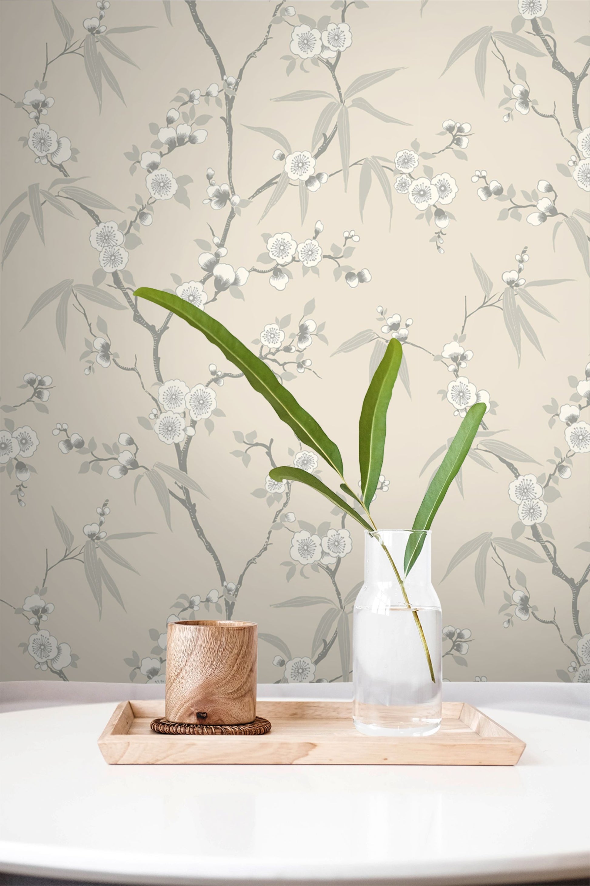 Vase with green plant on a wooden tray against a wall decorated with Beige & Soft Gray Blossom Spring Flower Wallpaper.
