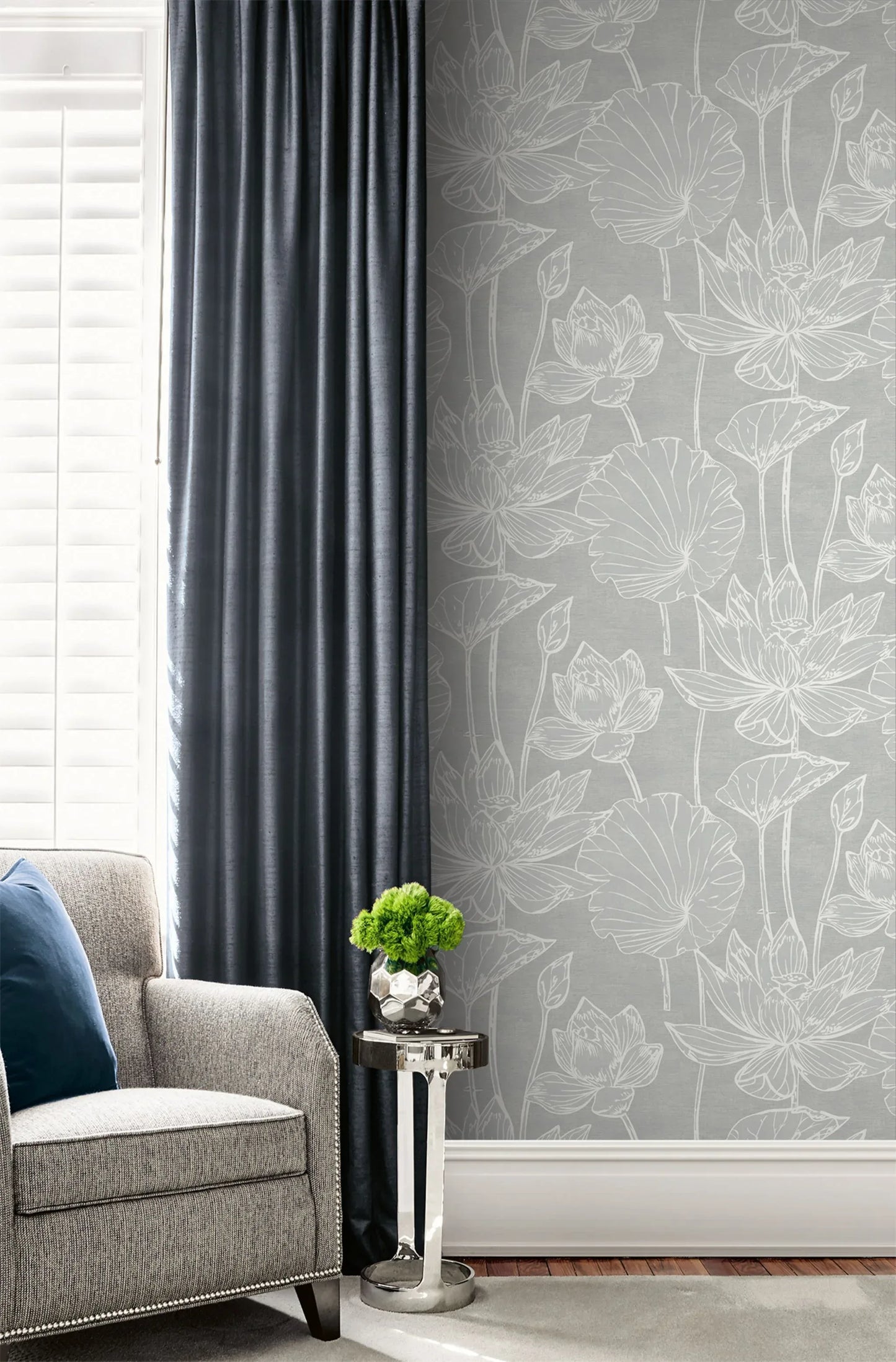 Gray armchair with blue pillow, small table, dark curtains, and Lily Flower Wallpaper near white shutters.