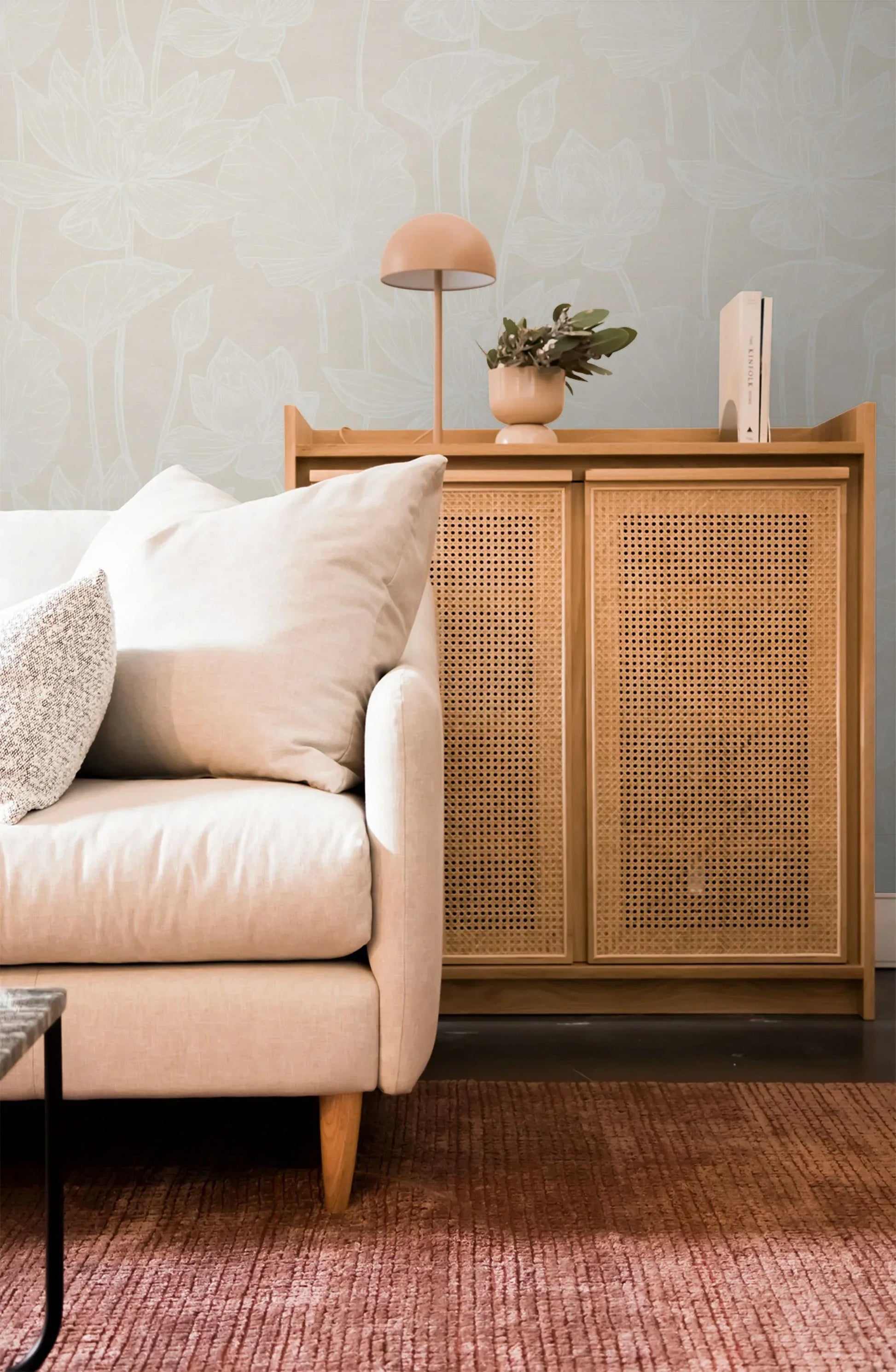 Light sofa beside a wooden cabinet with lamp and plant, on a pink rug with Lily Flower Wallpaper in the background.
