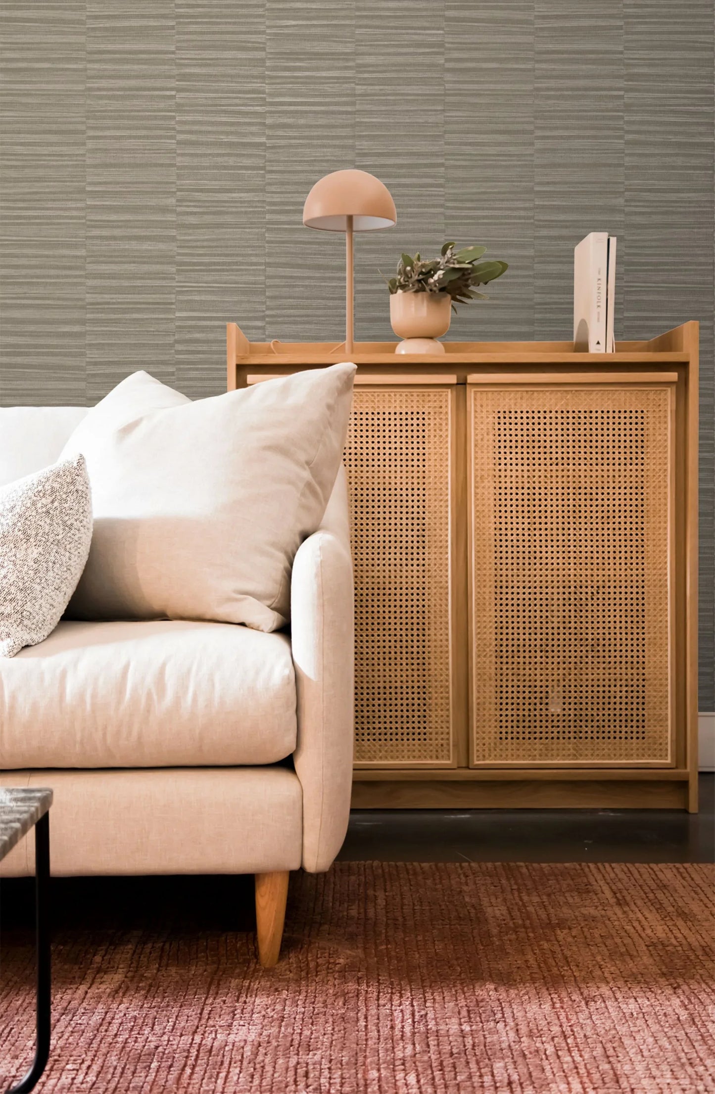 Beige sofa by wooden cabinet with plant, lamp, book, pink rug, and Industrial Grasscloth Stripe Vinyl wallpaper.